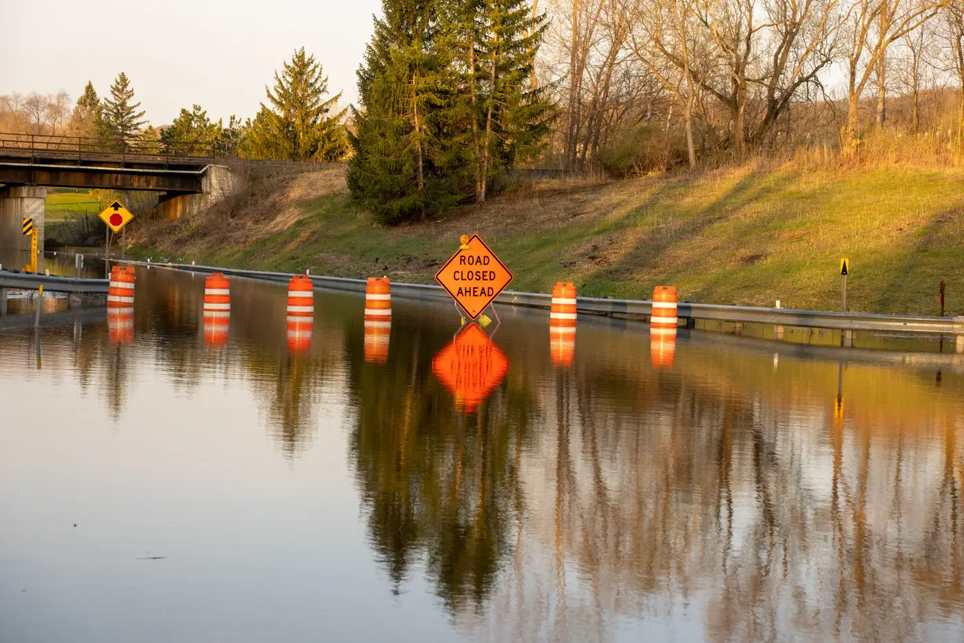 A flooded road leading to a bridge, lined with orange and white road-closed barricades, reflected in the still water.