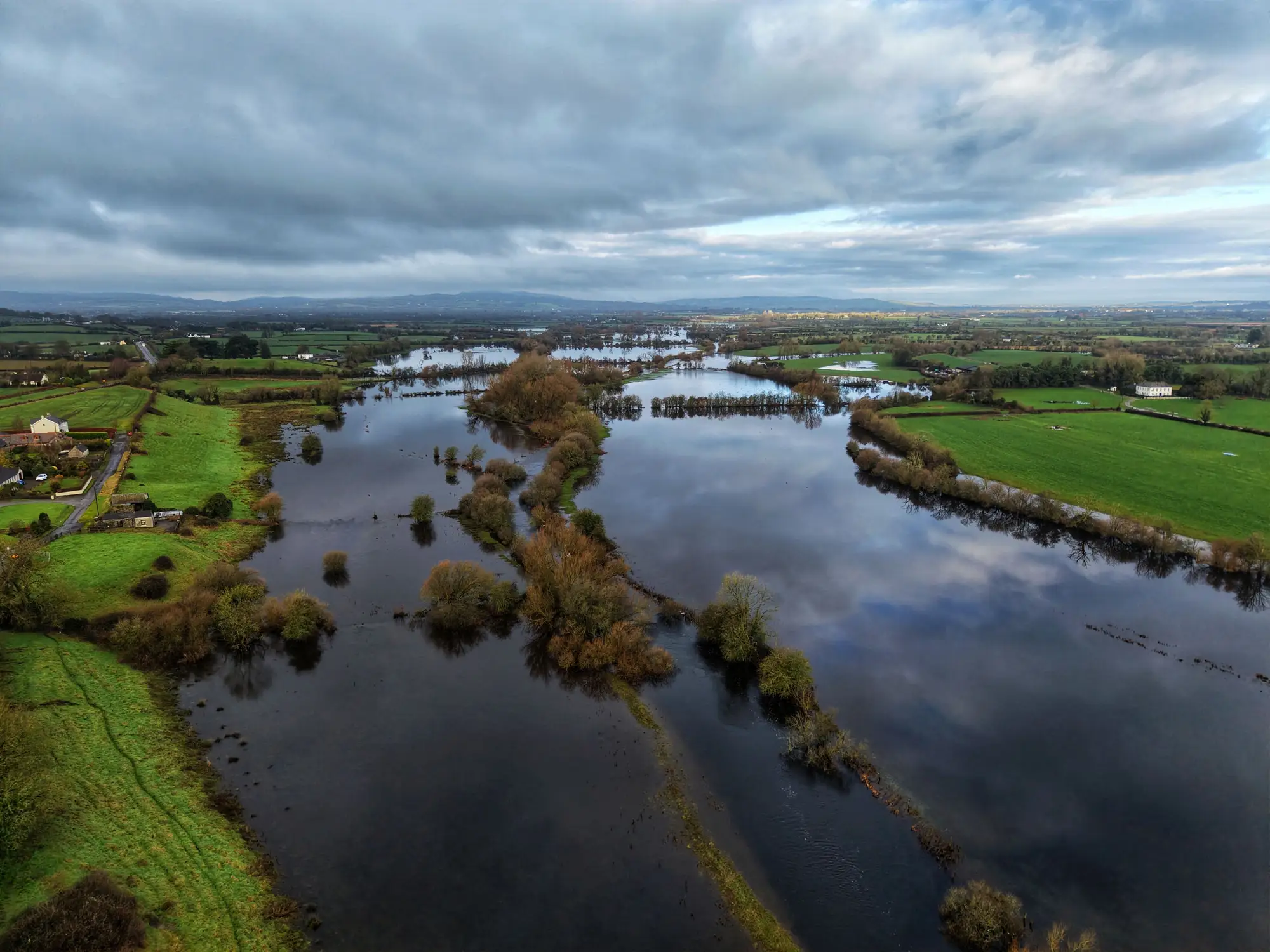 A river out of its banks across low fields under heavy cloud, seen from above the floodplain