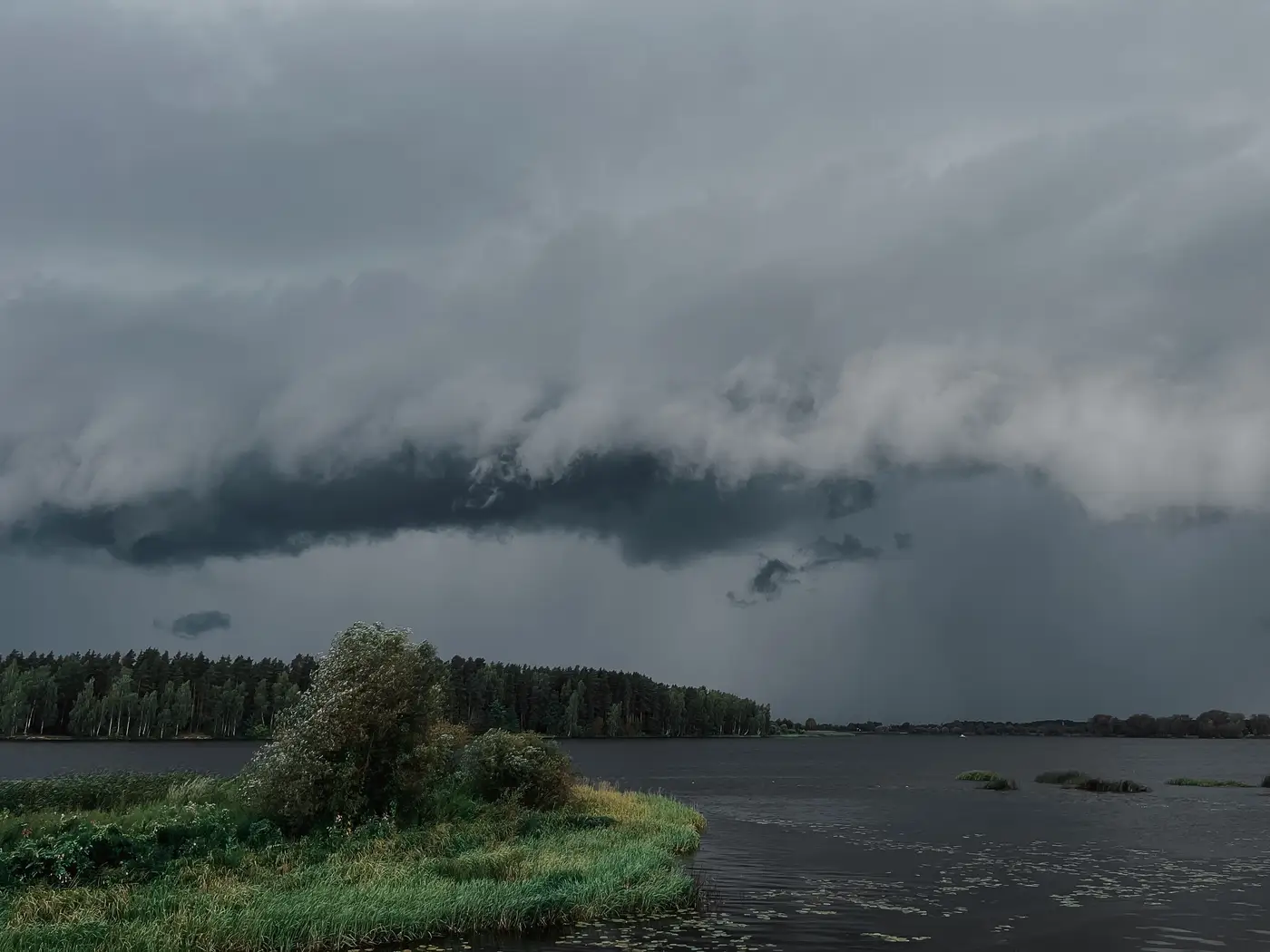 A dark storm cell looms over a lake and forested shoreline, rain visible falling in the distance.